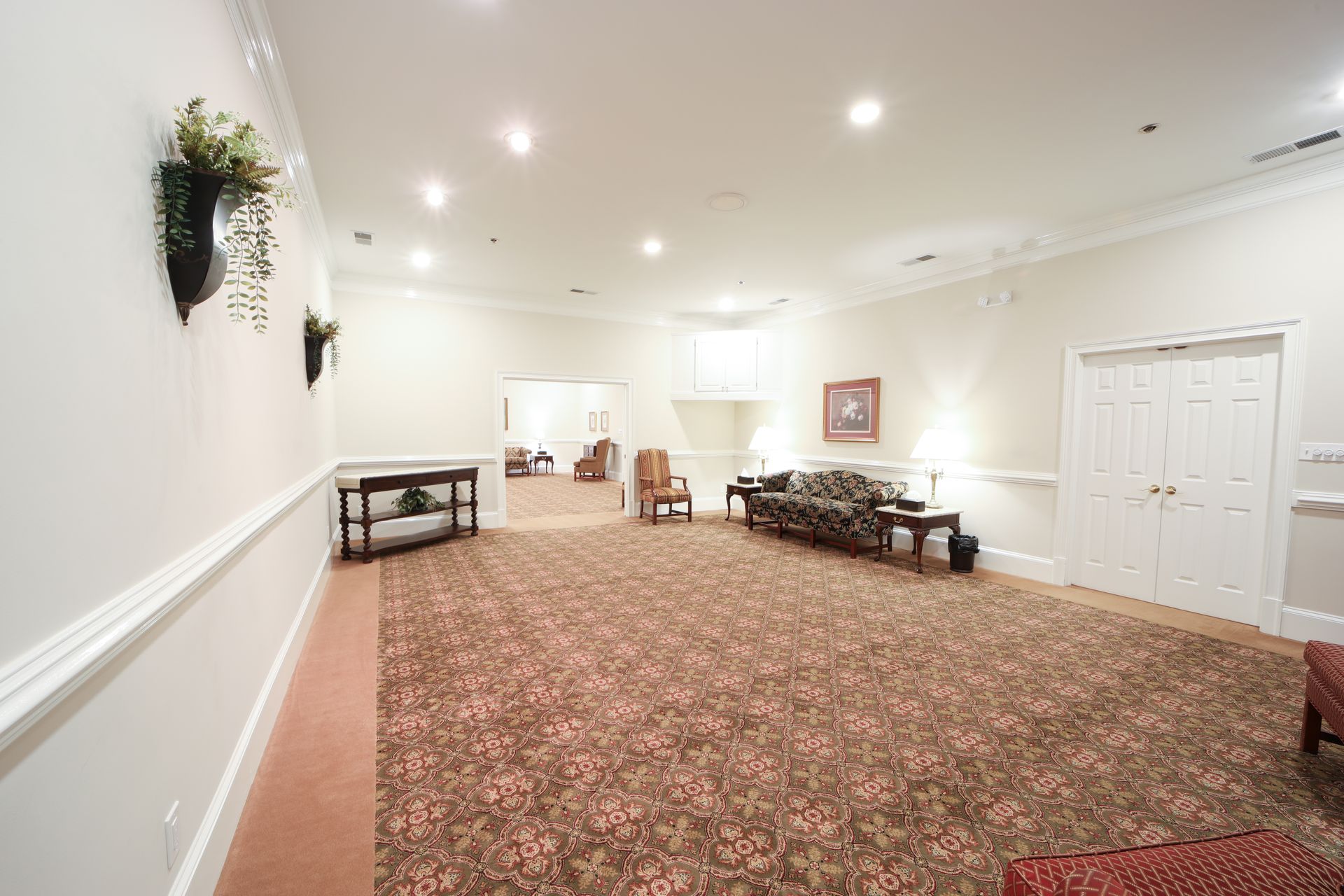 Large empty banquet hall with patterned carpet, white walls, recessed lights, tables, and double doors