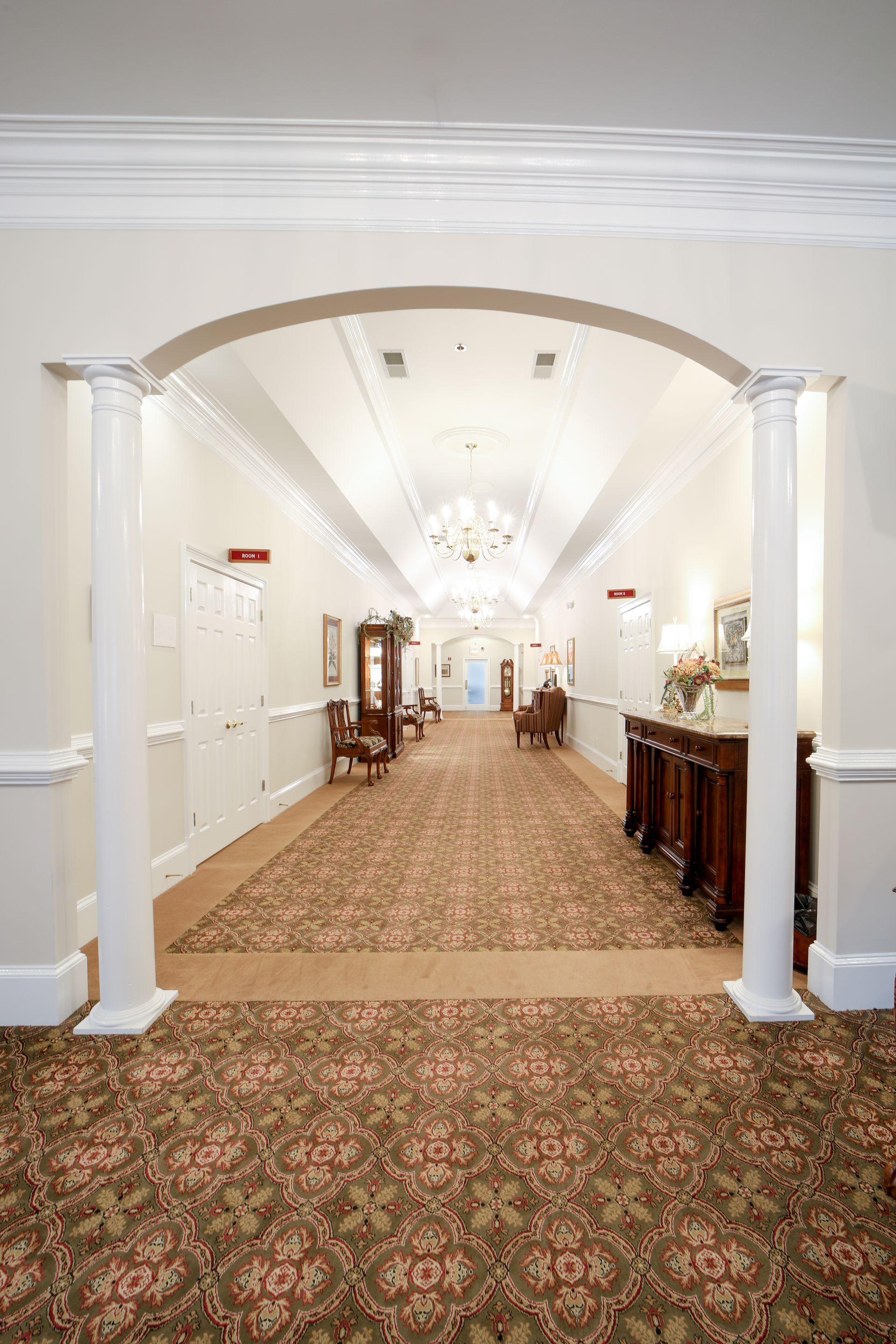 Elegant hotel hallway with patterned carpet, white columns, and antique furniture leading to a bright chandelier-lit room