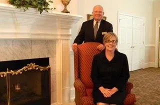 Two people posing in a white-paneled room with a fireplace and red patterned chair