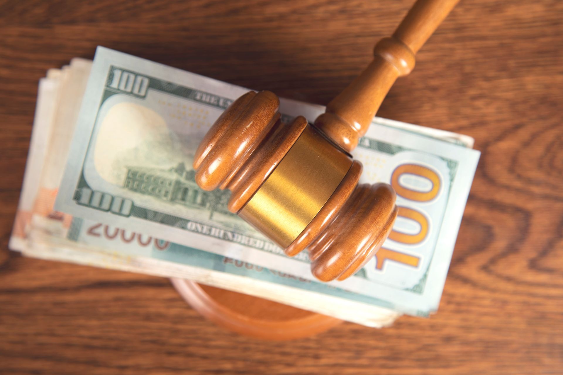 A wooden gavel resting on a stack of US 100-dollar bills on a brown wooden surface.