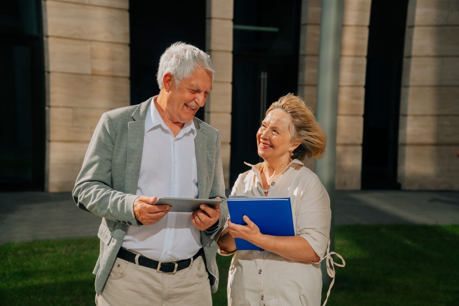 Two people smiling and looking at a tablet outside a building, one holding a blue folder