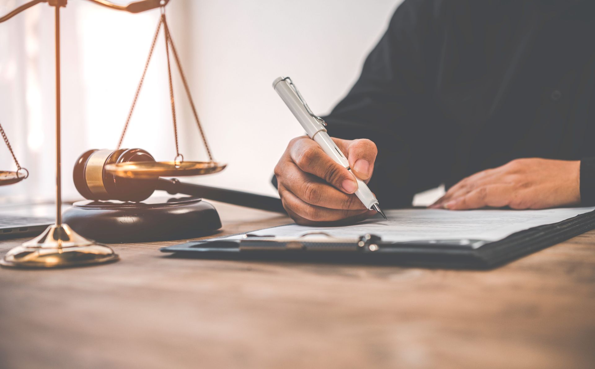 A person writes on a clipboard next to a golden gavel and a scale of justice on a wooden desk.