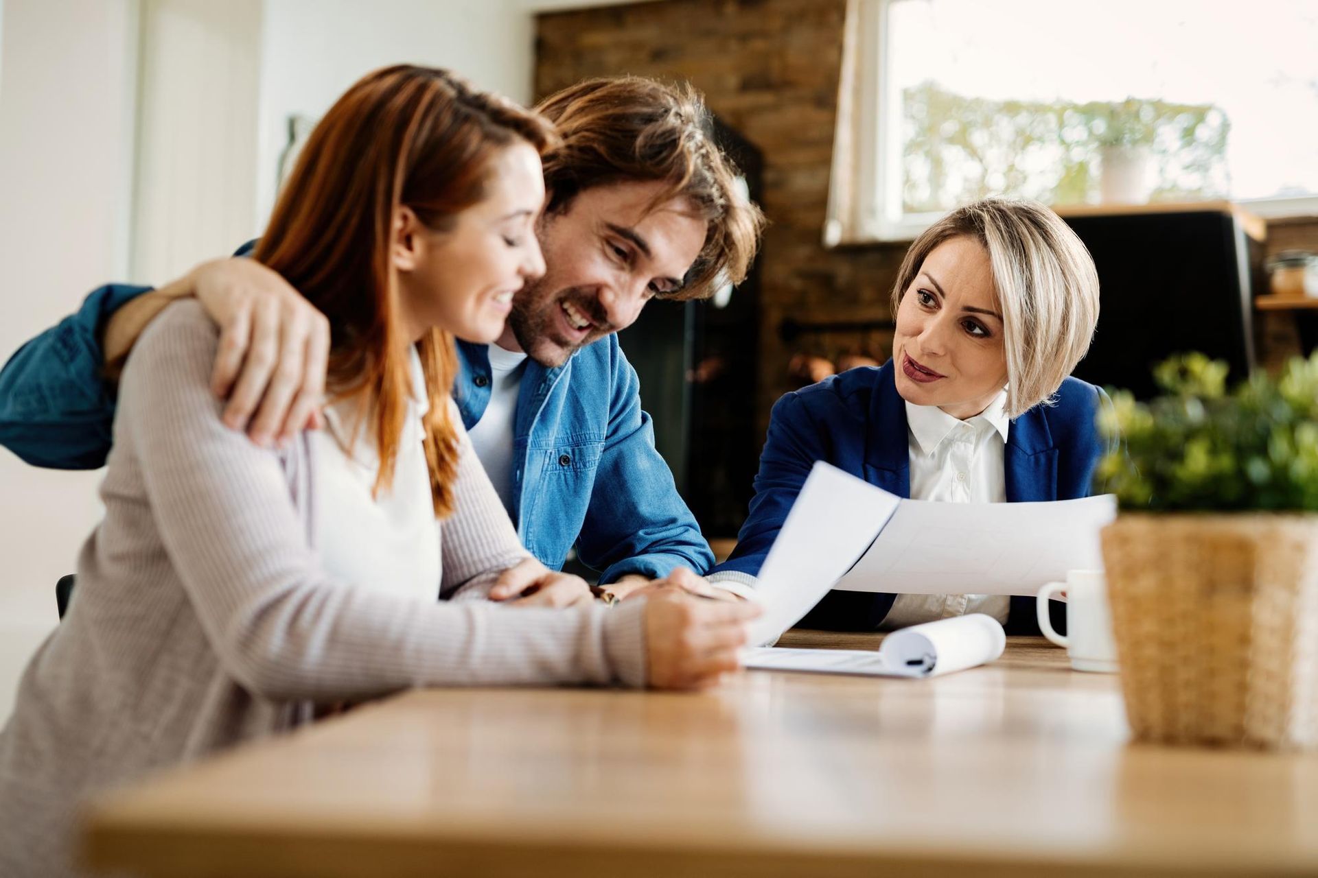 A couple sits at a wooden table reviewing documents with an advisor, who is pointing at a paper.