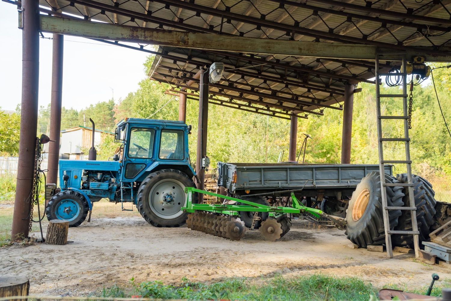 A blue tractor is parked under a shed next to a gray trailer, a green harrow, and two large spare tires.
