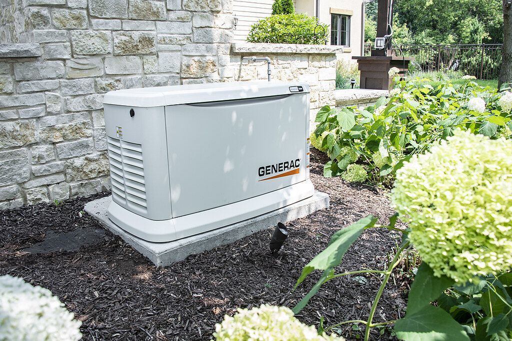 Outdoor generator beside a stone house, surrounded by mulch and white flowers