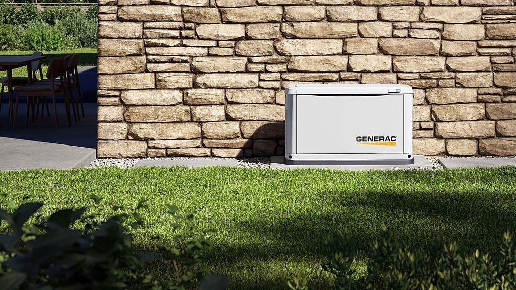 White outdoor generator beside a stone wall, with grass in the foreground.