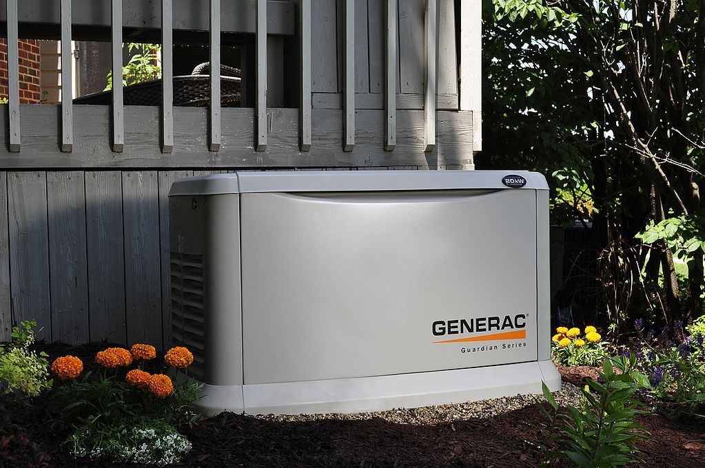 Gray Generac standby generator beside a house, surrounded by flowers and mulch.