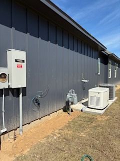 Side of a gray building with utility boxes, air conditioners, and exposed equipment on dry grass.