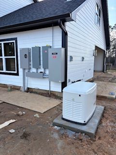 Outdoor home utility equipment beside a house, including gray electrical boxes and a white generator on a concrete pad