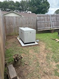 Outdoor air conditioner unit on a concrete pad in a fenced backyard, with grass and a tree stump nearby.