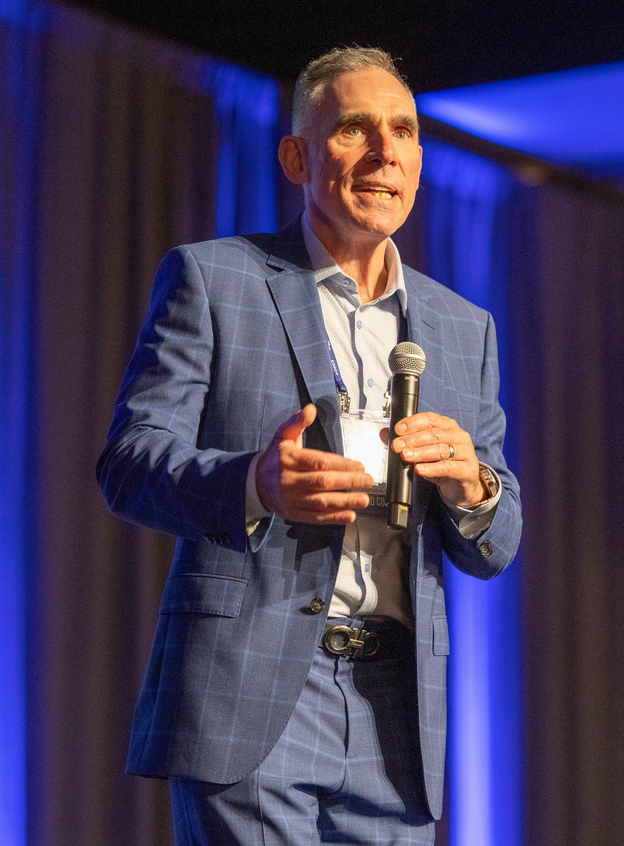 Man in a blue suit speaking on stage, holding a microphone under purple lighting.