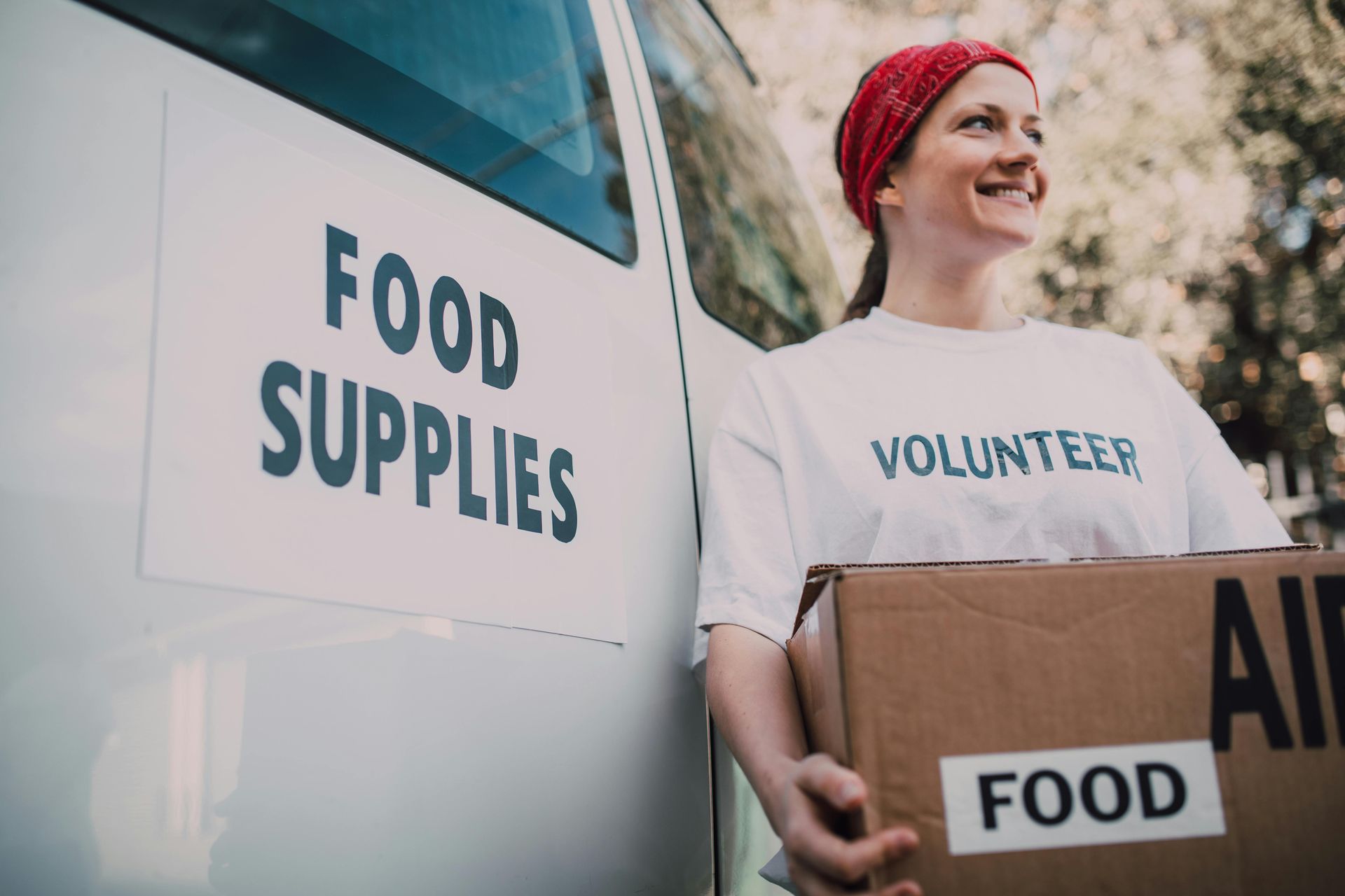 Volunteer holding food boxes beside a food supplies truck, wearing a red knit hat and smiling