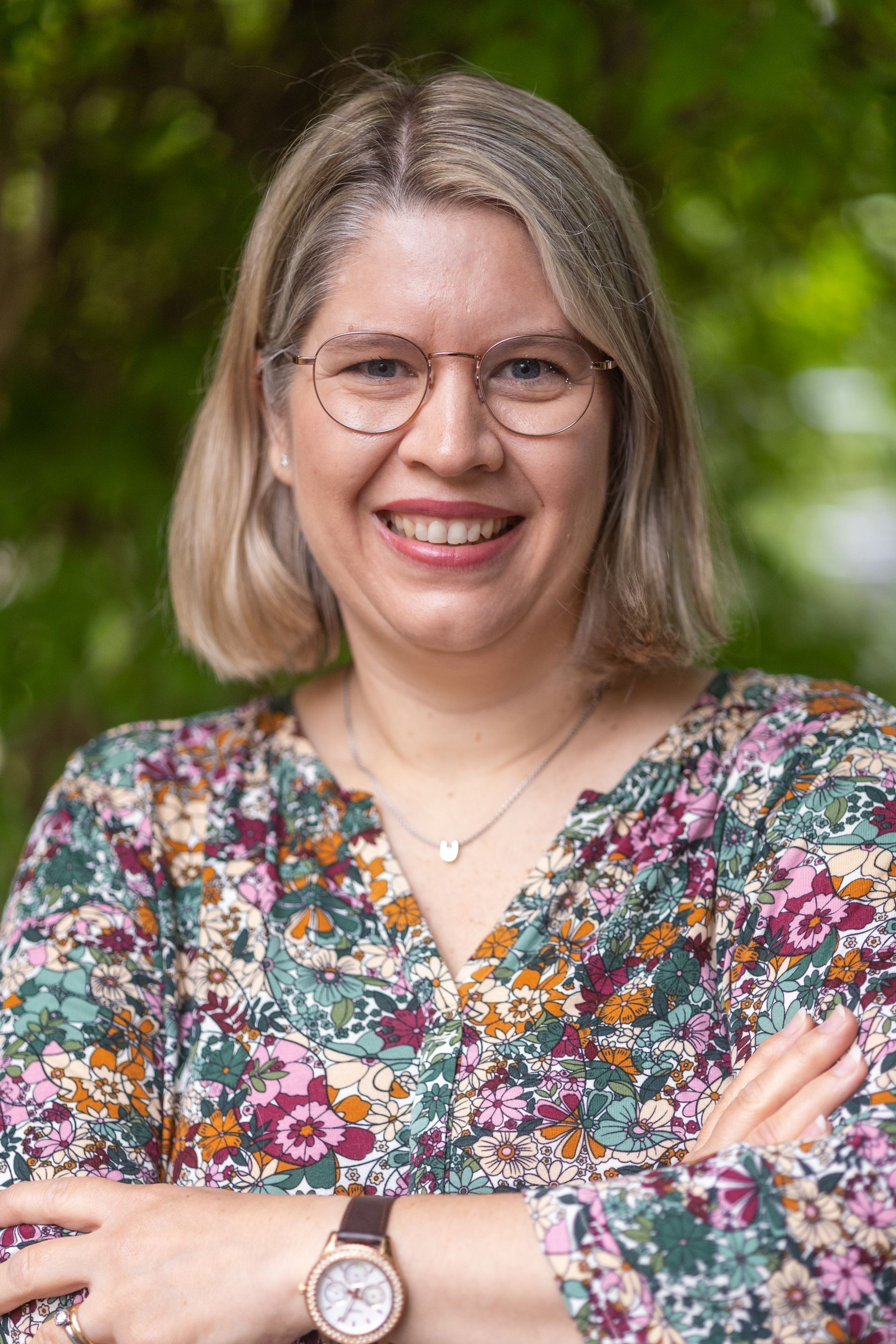 A woman wearing glasses and a floral shirt is smiling with her arms crossed.