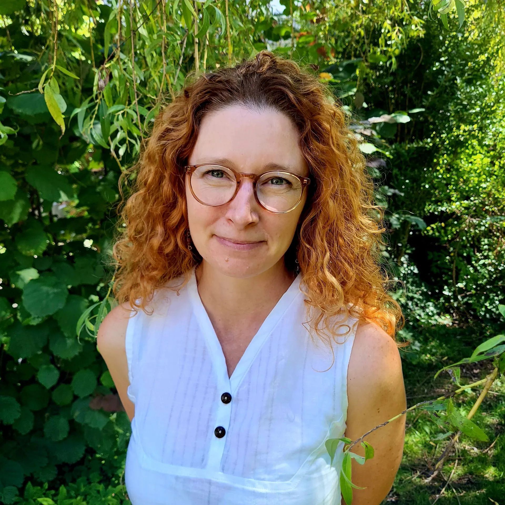 A woman with red hair and glasses is standing in front of a bush.