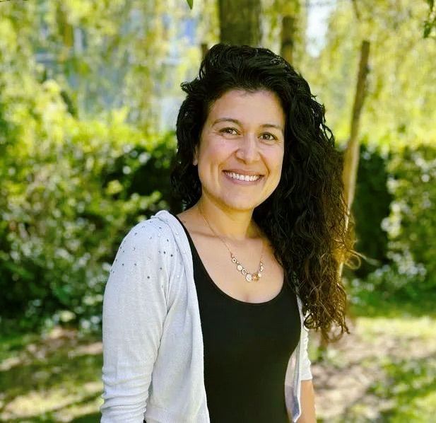 A woman in a black dress and white cardigan is smiling in a park.