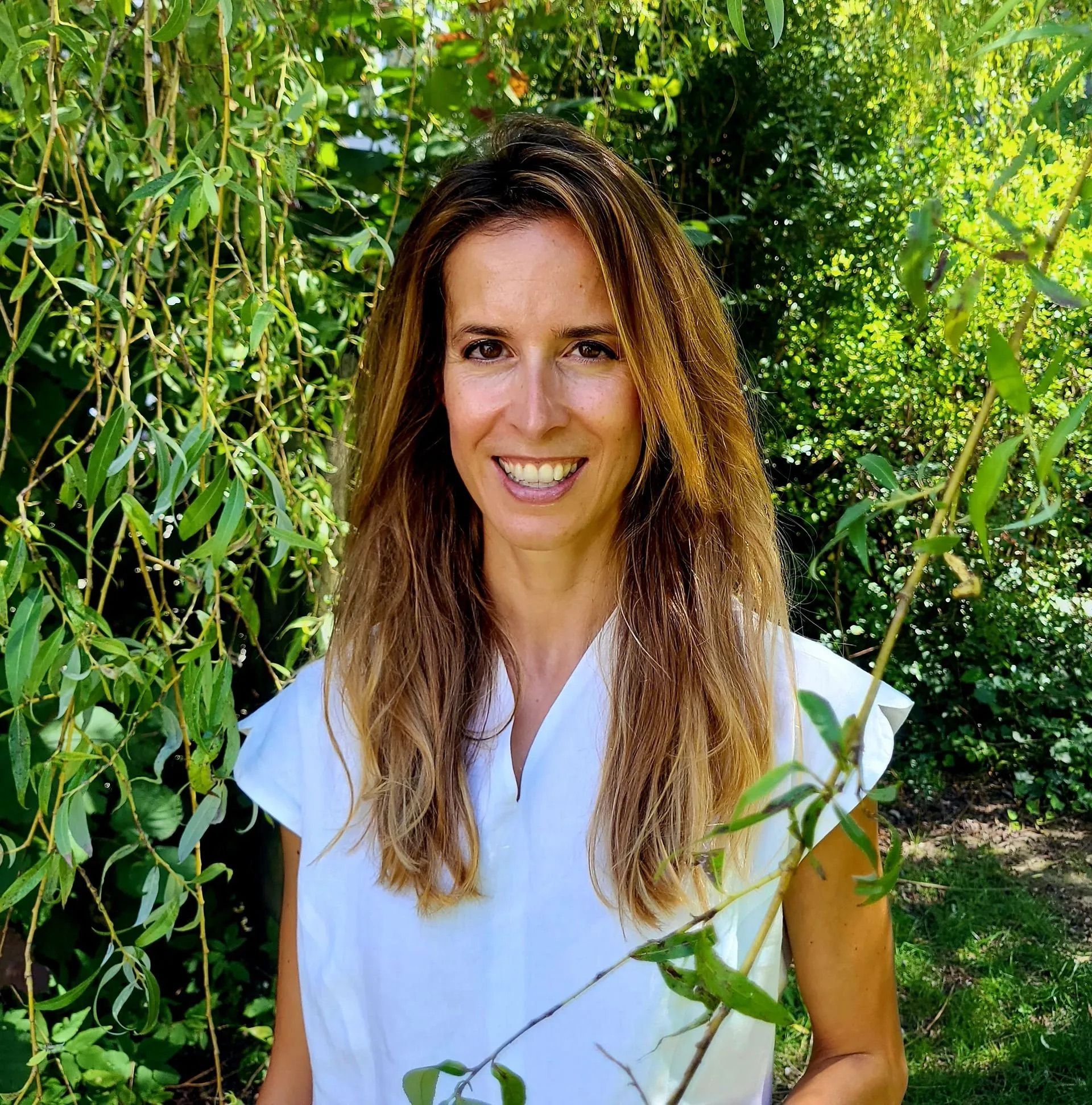A woman in a white shirt is smiling in front of a tree.