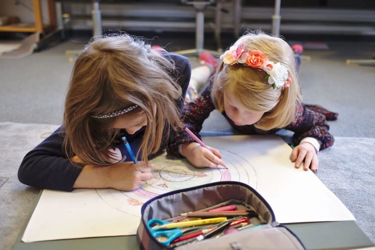 Two young girls are laying on the floor drawing on a piece of paper.