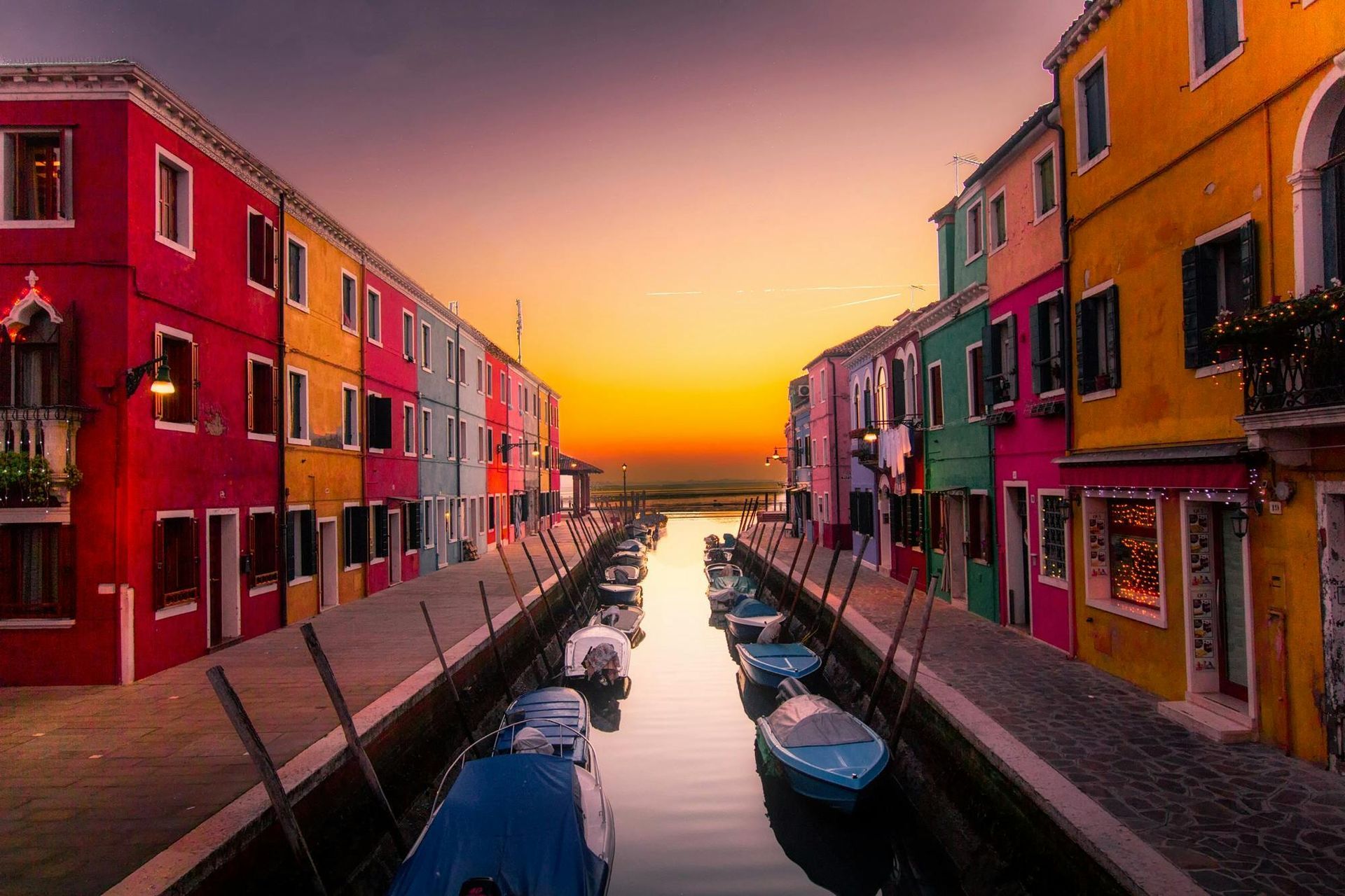 A row of colorful buildings along a canal with boats in it at sunset.