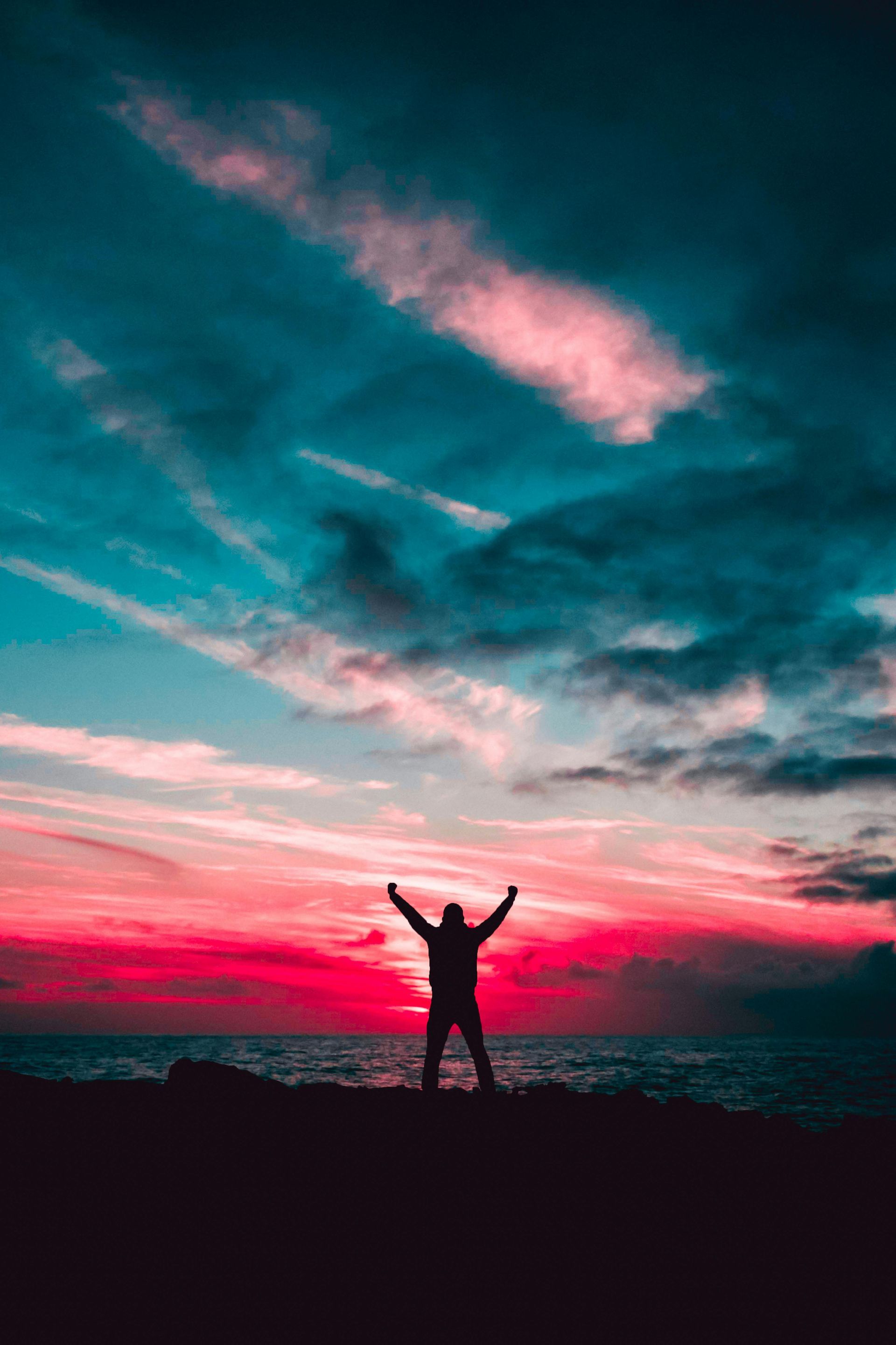 A man is standing on a beach with his arms in the air at sunset.