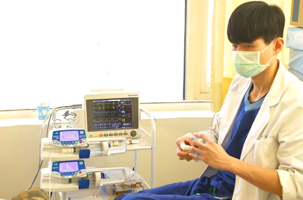 A doctor wearing a mask is sitting in front of a monitor in a hospital room.