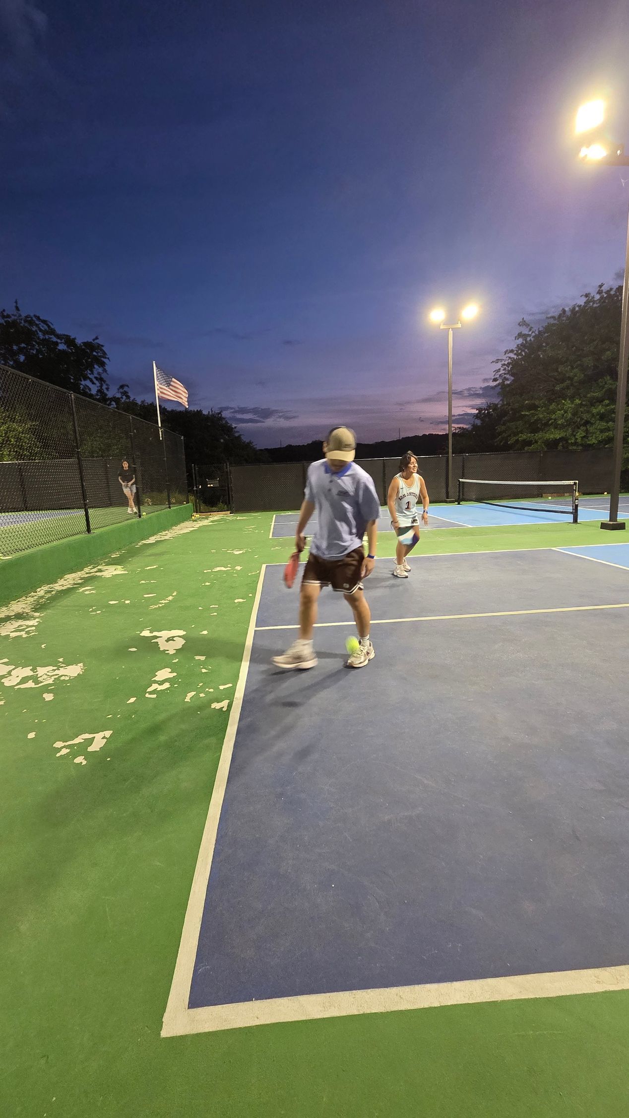 People on an outdoor tennis court at dusk, with blue and green painted surfaces and lights overhead.
