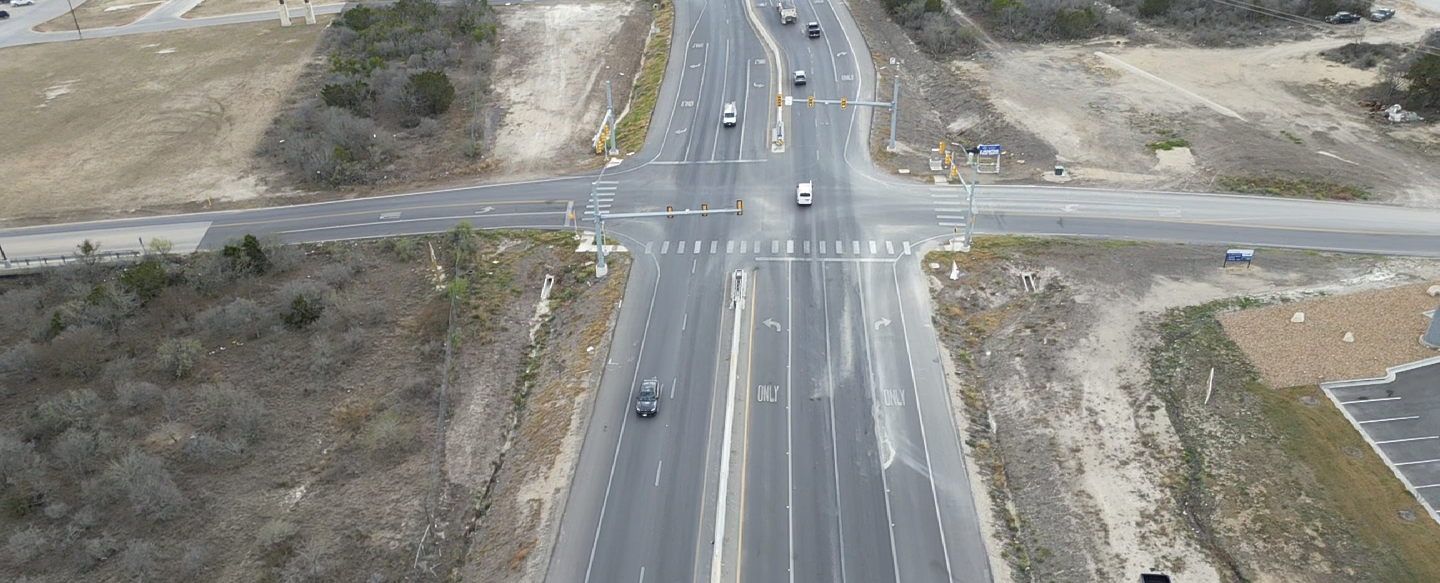 Aerial view of a road intersecting. Cars travel on asphalt, surrounded by green trees and homes.
