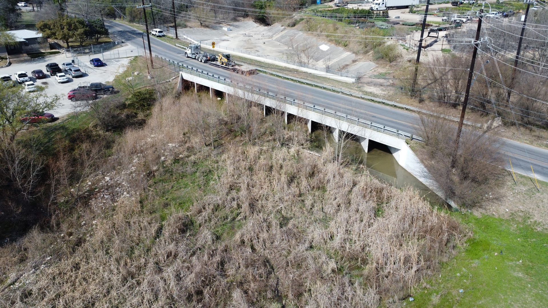 Aerial view of a concrete bridge over a steep, grassy embankment beside a road and parking lot.