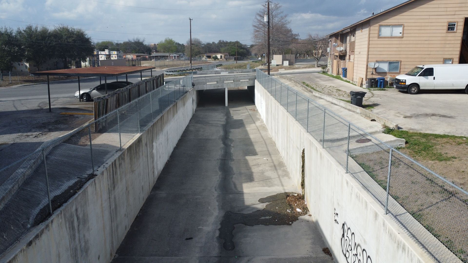 Empty concrete drainage channel running between retaining walls beside a building and parking area.