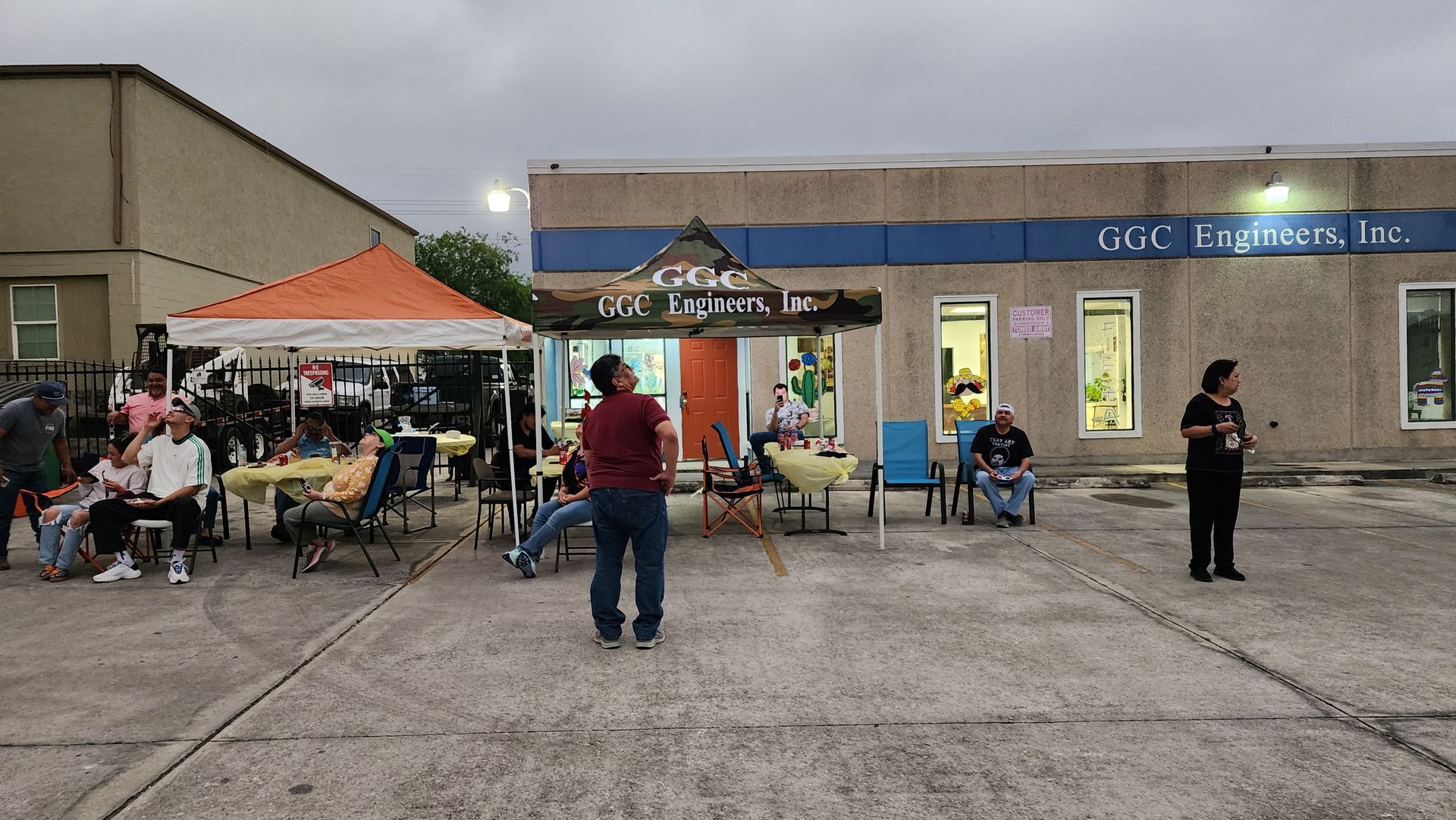 Outdoor community gathering under tents in a parking lot, with people seated and standing near a brick building.