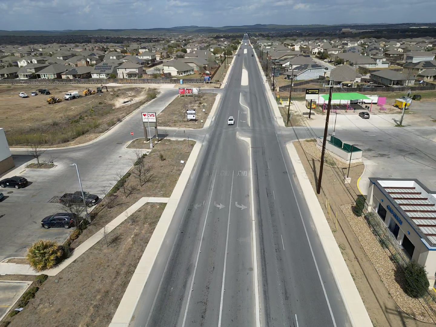 Aerial view of a long straight road through a suburban neighborhood with scattered houses and open lots.