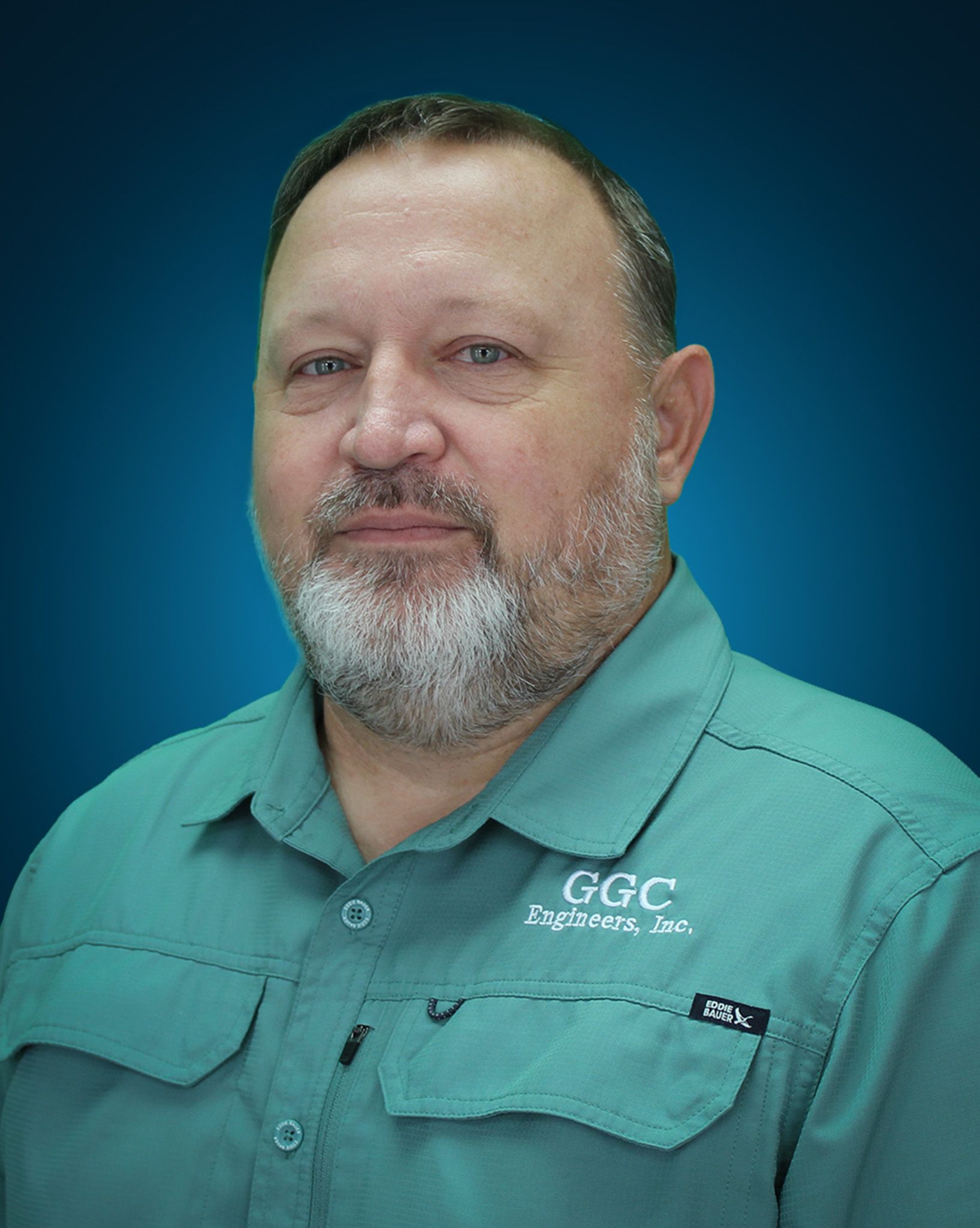 Man in teal shirt, looking at camera. Gray beard, blue background, 