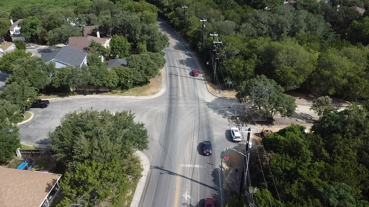 Aerial view of a road intersecting. Cars travel on asphalt, surrounded by green trees and homes.