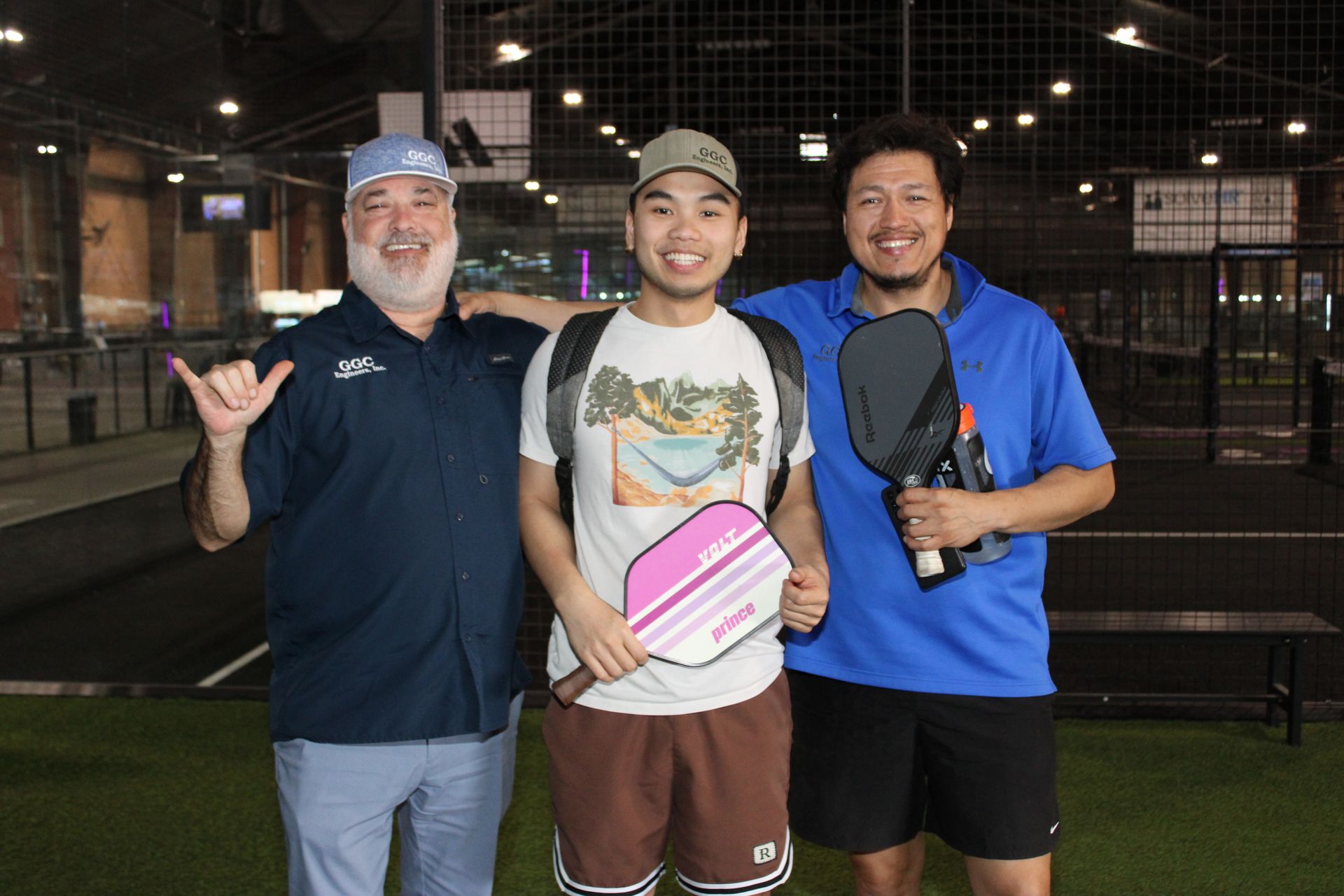 Three GGC Engineering team members holding pickleball paddles pose indoors on a court with a net behind them.