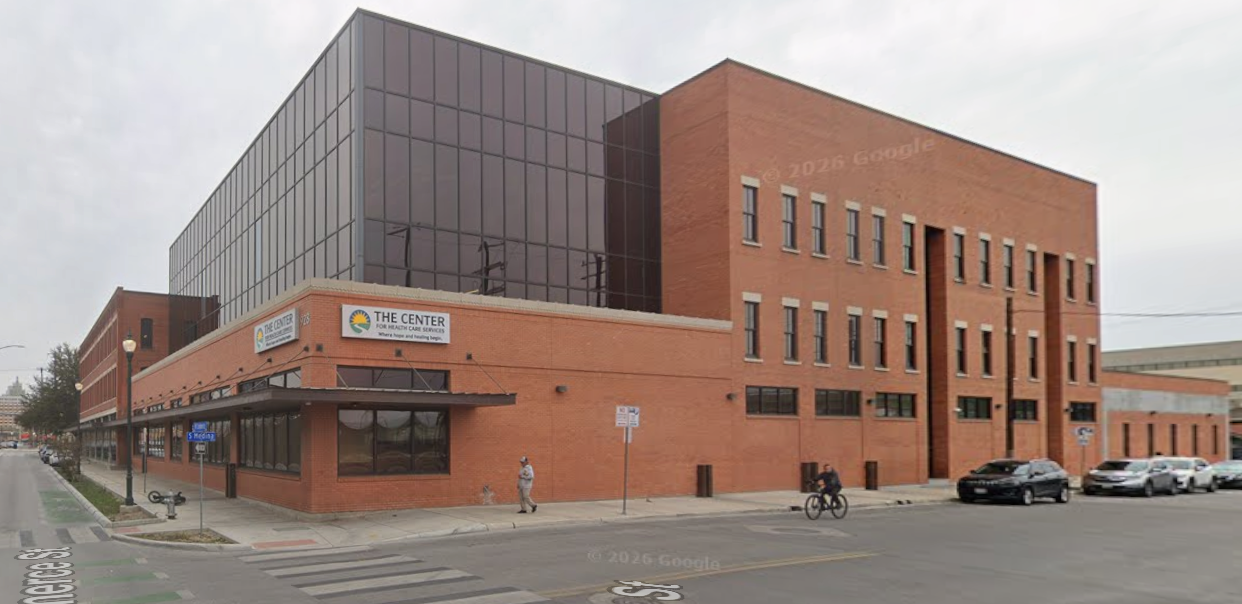 Brick commercial building on a city street corner with large glass windows and a sidewalk storefront.