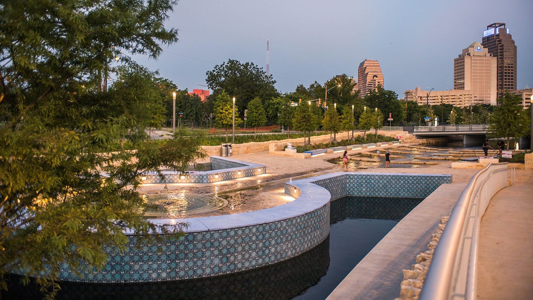 Curved mosaic fountain and reflecting pool in a park, with trees and city buildings in the background