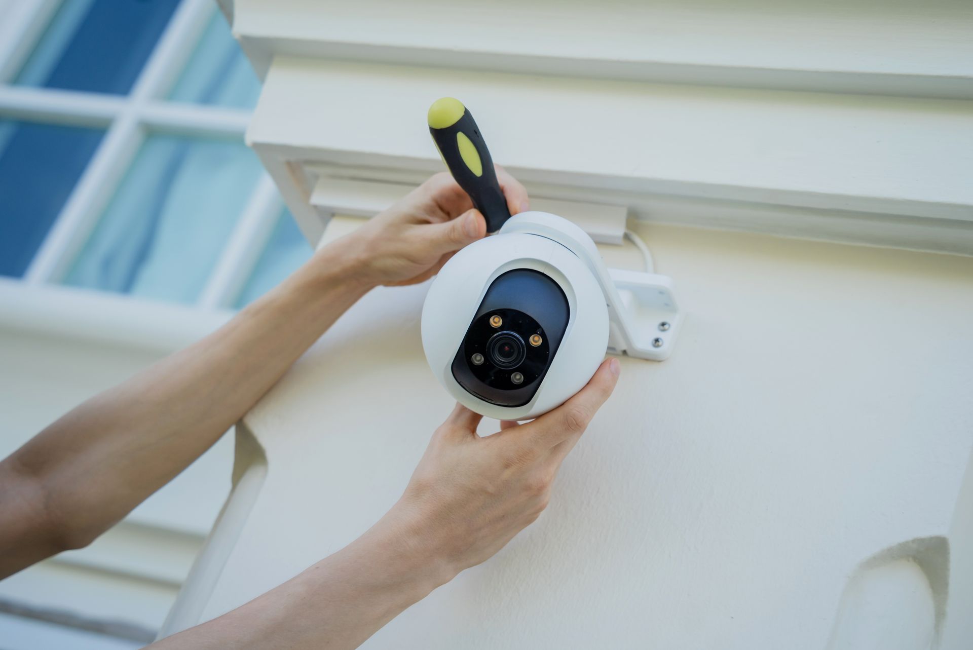 A person uses a screwdriver to mount a white, dome-shaped security camera onto a light-colored exterior wall.