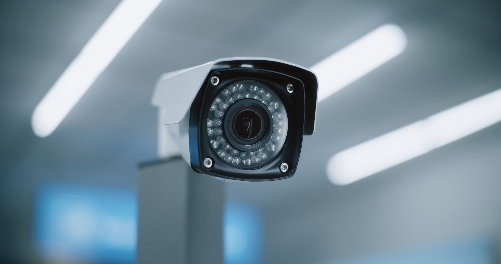 A modern white security camera mounted on a pole against a blurry, light-colored indoor background with overhead lights.