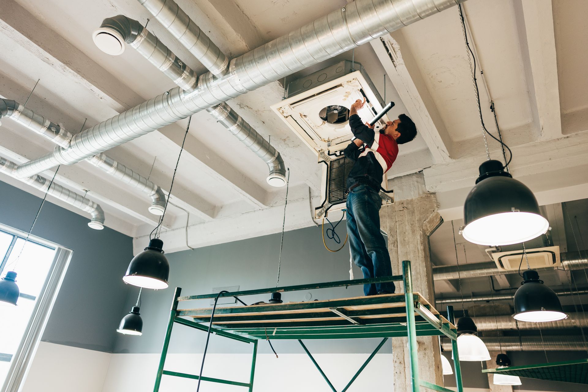 A technician on scaffolding works on an industrial ceiling-mounted air conditioning unit in a modern space.