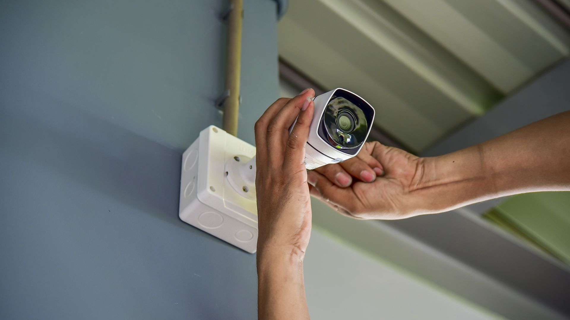 Hands adjusting a white security camera mounted on a gray wall next to a junction box.