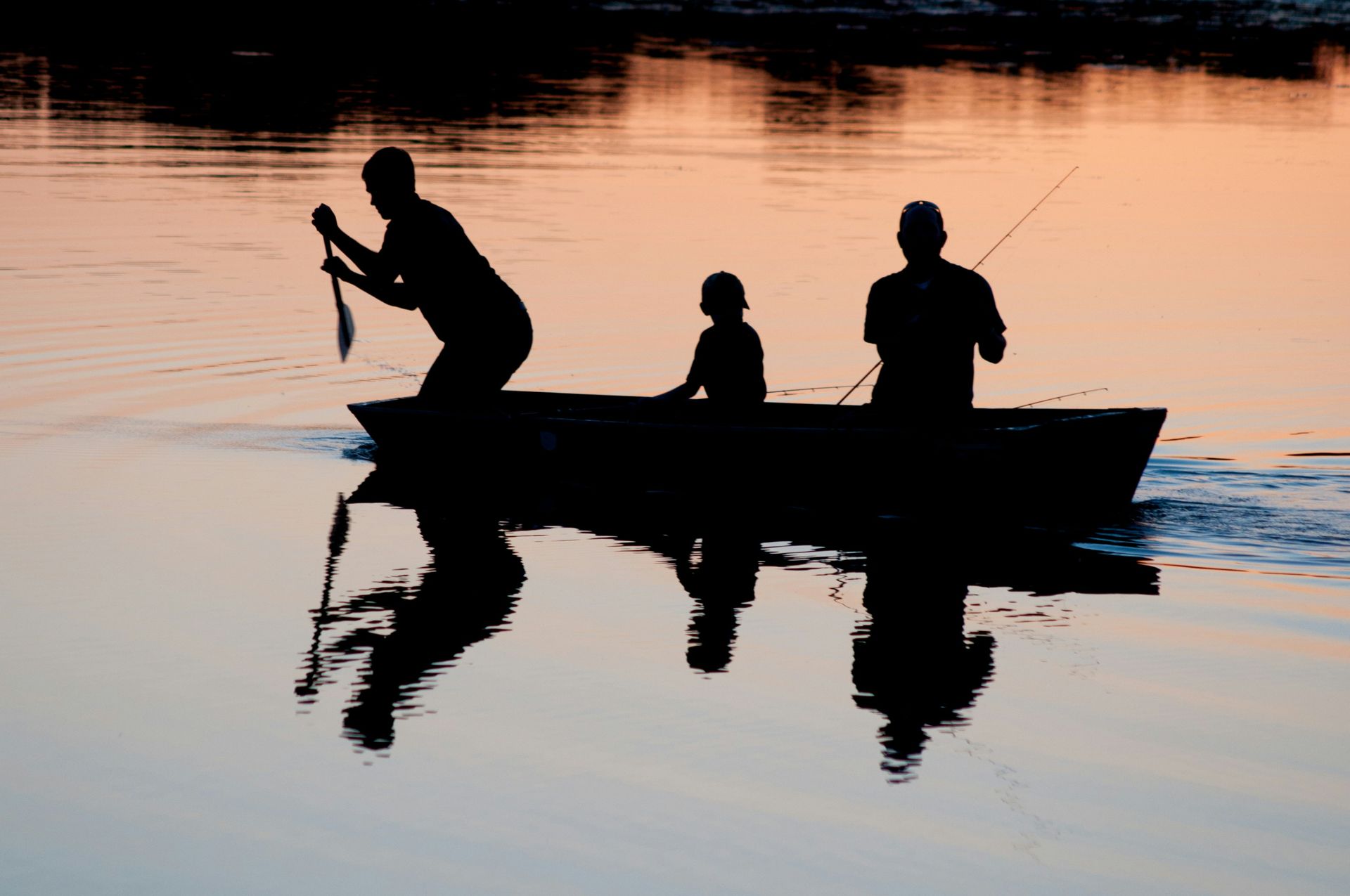Three people in a boat are fishing in the water at sunset