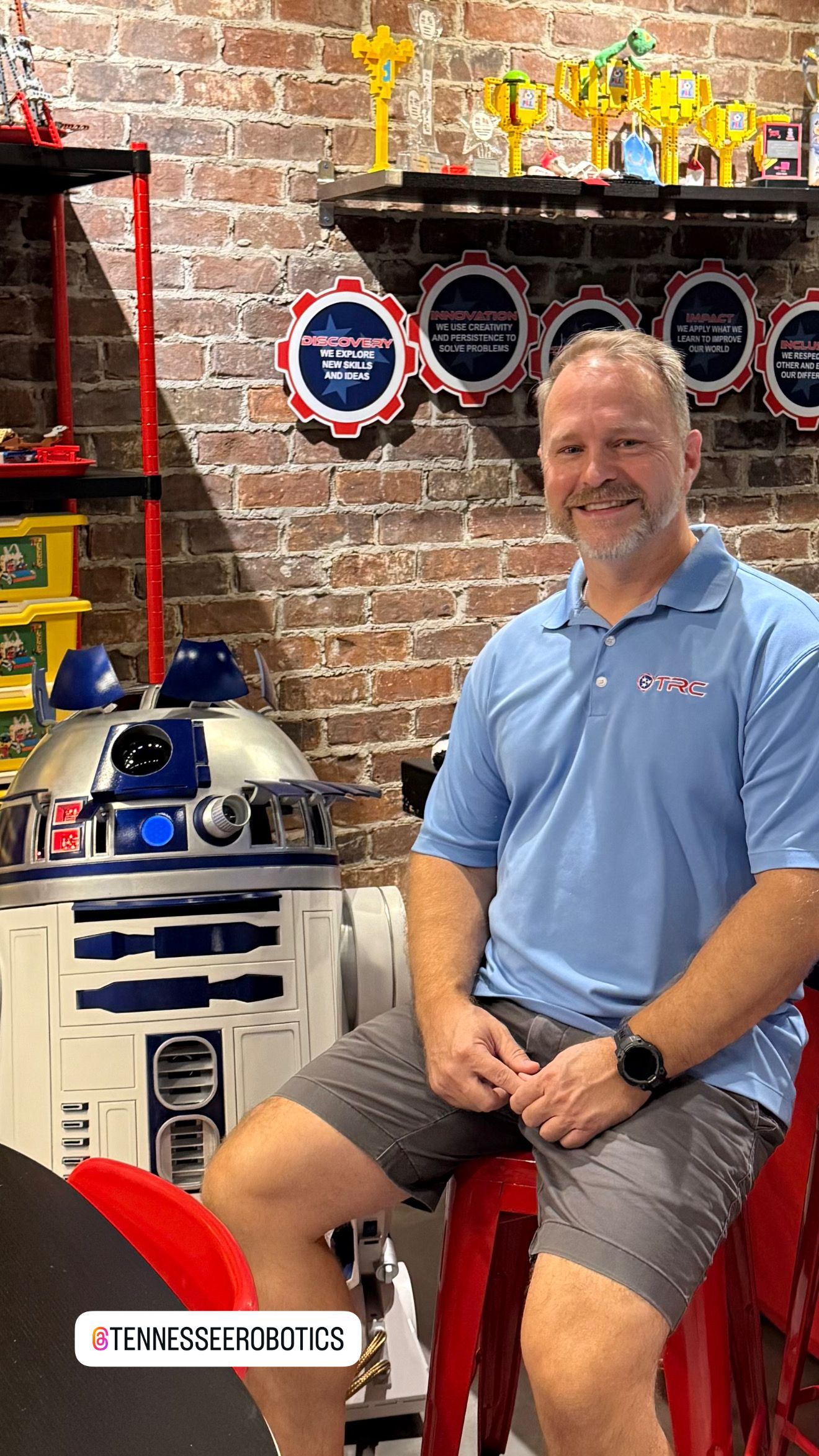 Man seated with a R2-D2 robot, near brick wall.  Shelves with trophies and a sign. Man in blue shirt, smiling.
