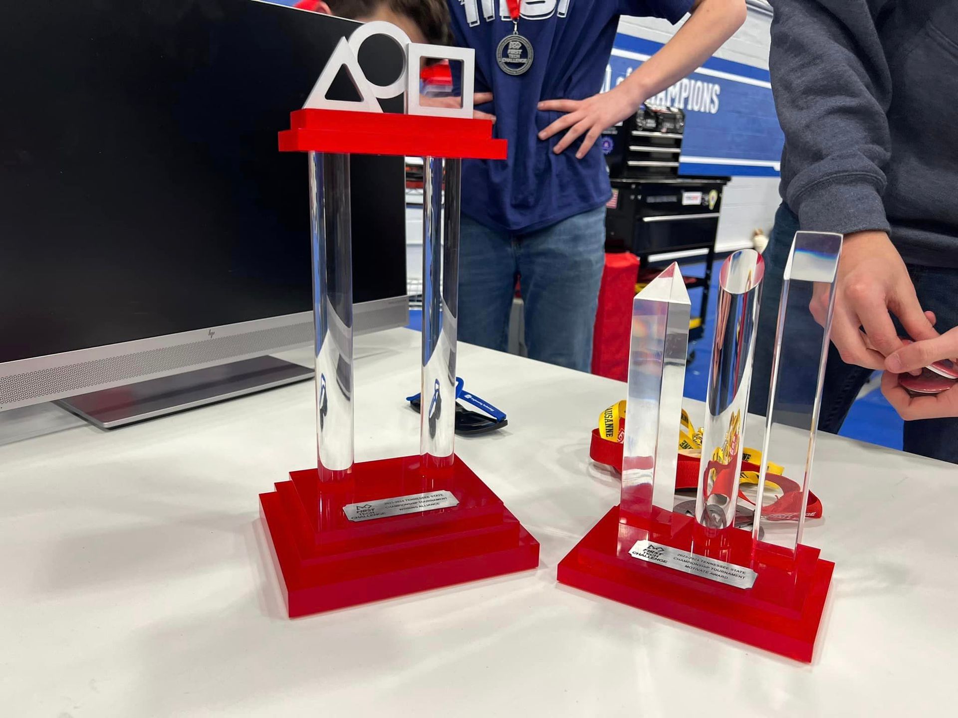 Two trophies on a table with red bases and clear pillars. A person wearing a medal stands nearby.
