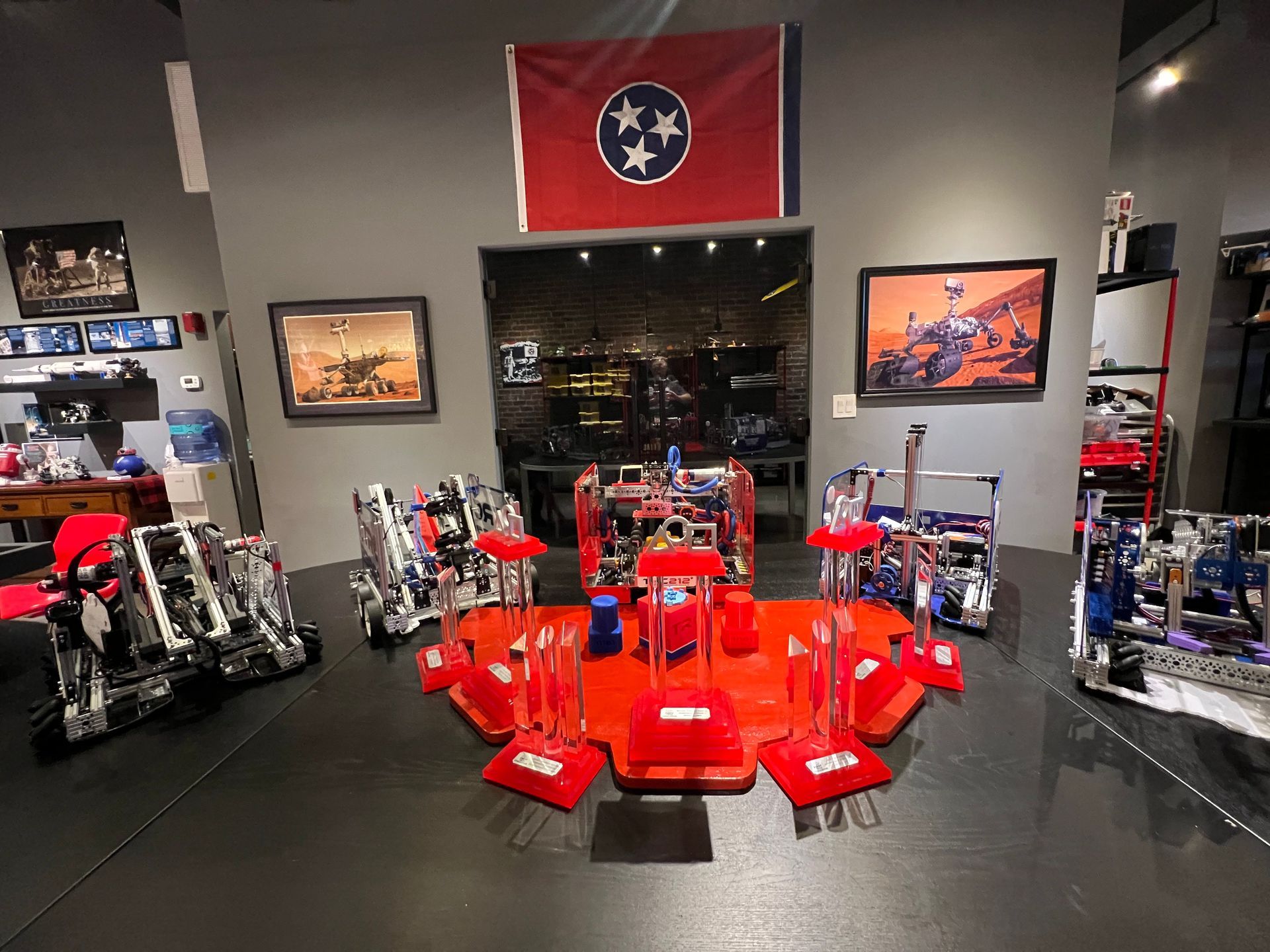 Robotics display: red & silver robots on a black floor, under a TN flag and space art.