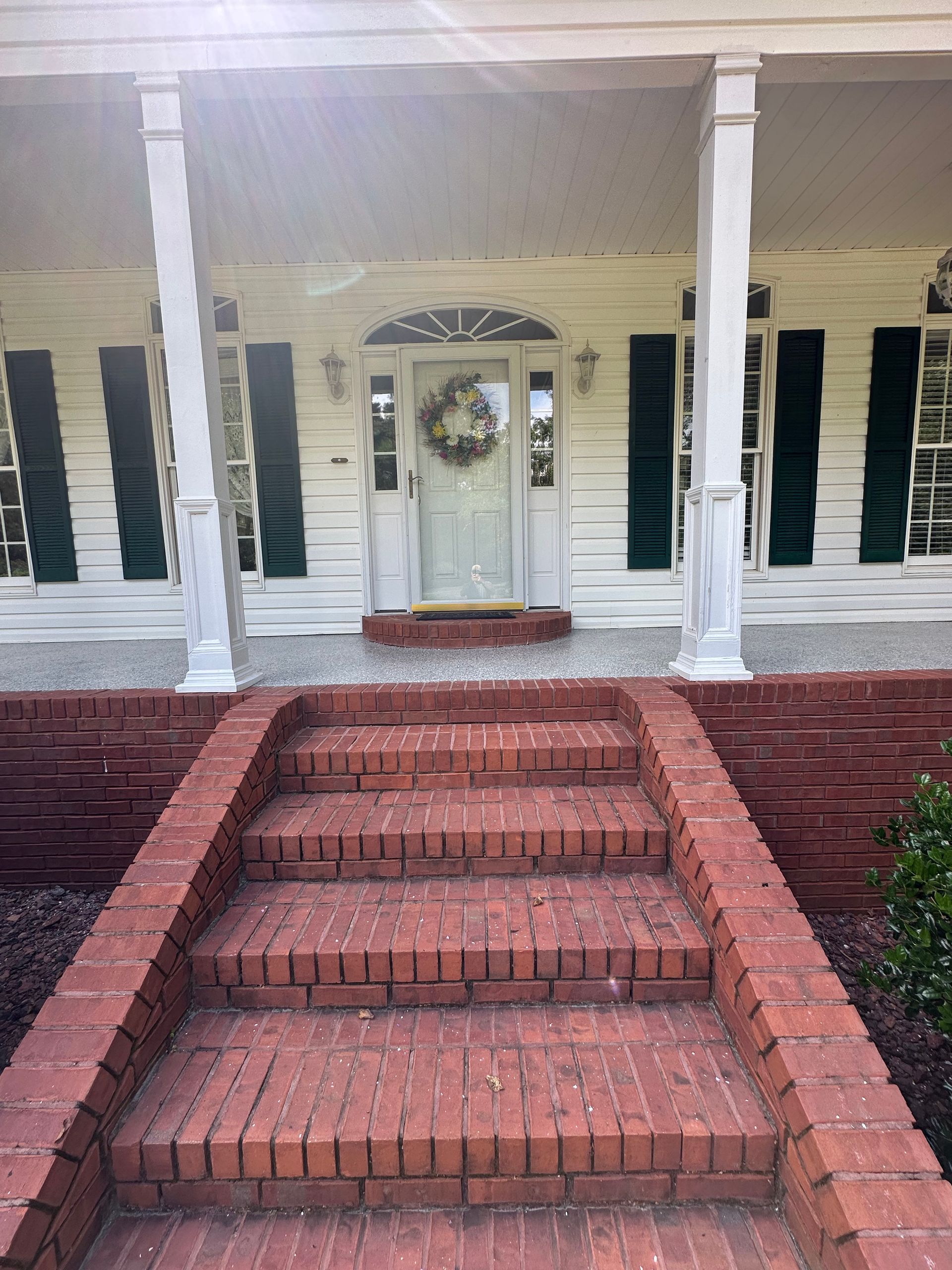 Brick steps leading to a white house porch with green shutters and a light-colored front door decorated with a wreath.