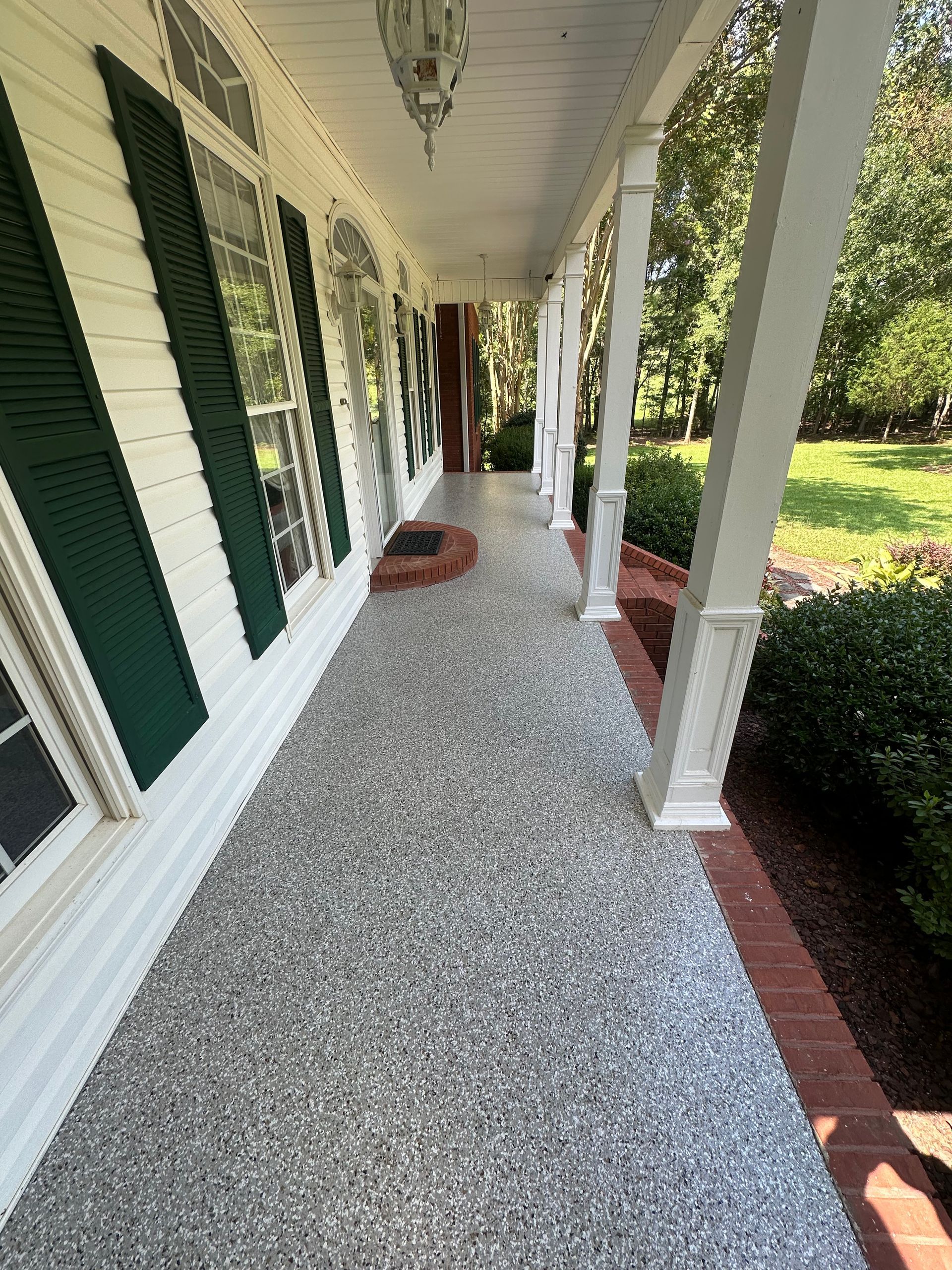 A long, covered residential porch with light yellow siding, green shutters, and a speckled gray composite floor.