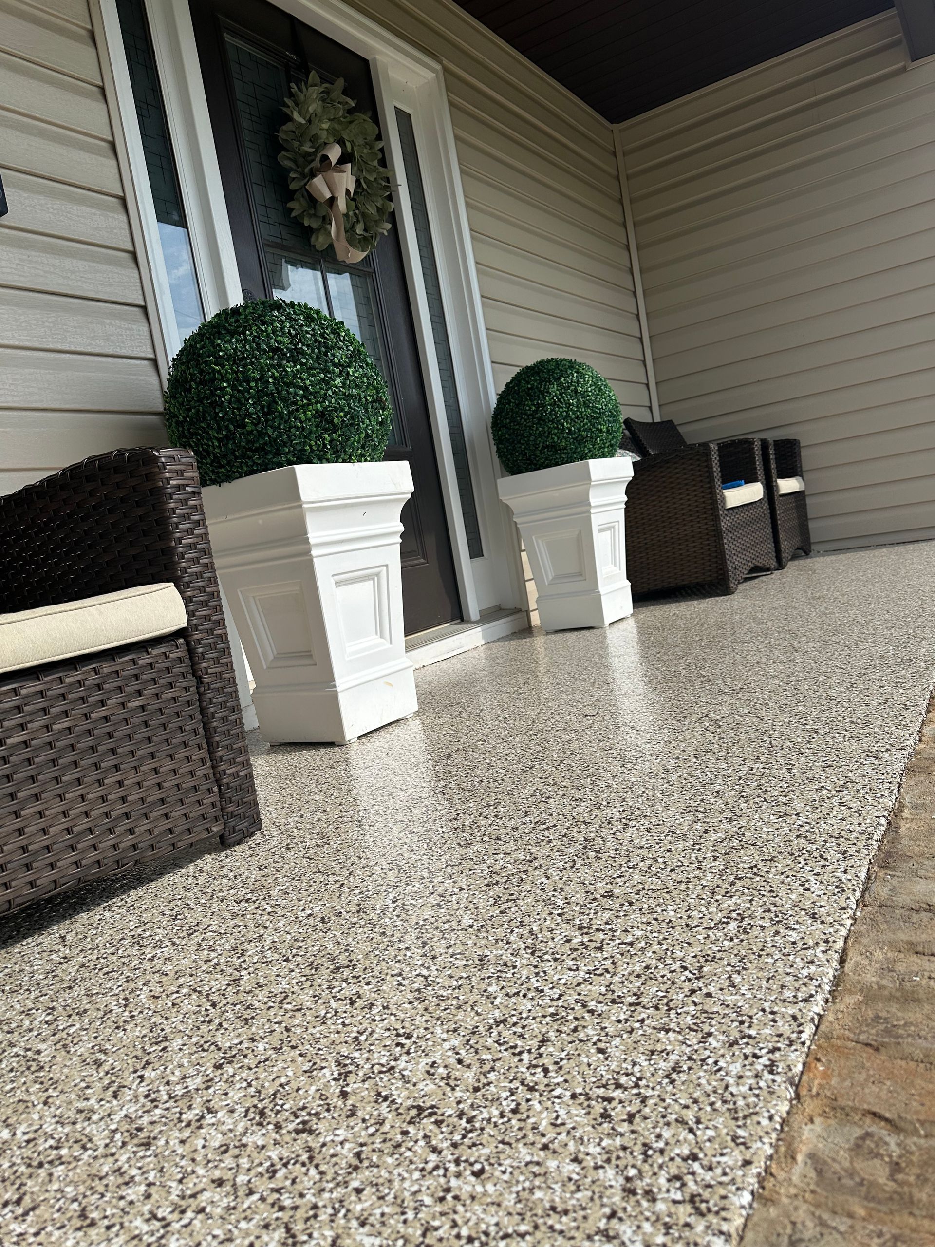 Porch with a speckled epoxy floor, two brown wicker chairs, and topiary plants in white square planters.