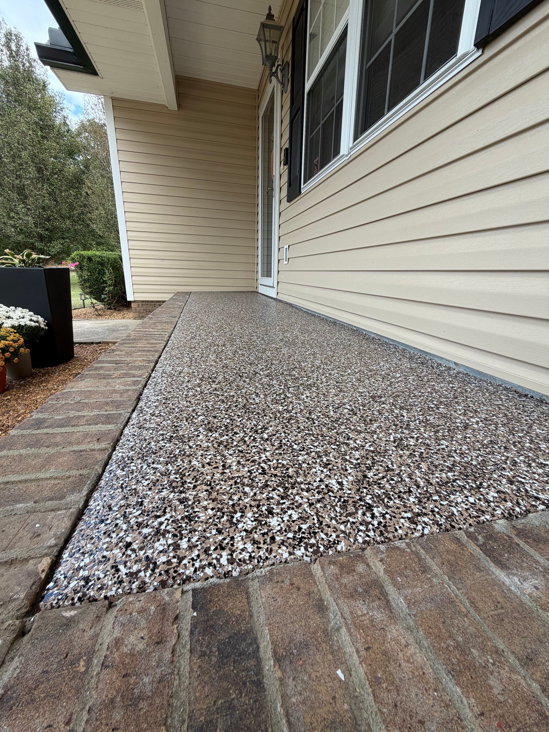 A porch floor with a textured, speckled epoxy coating, bordered by bricks, next to a beige-sided house exterior.