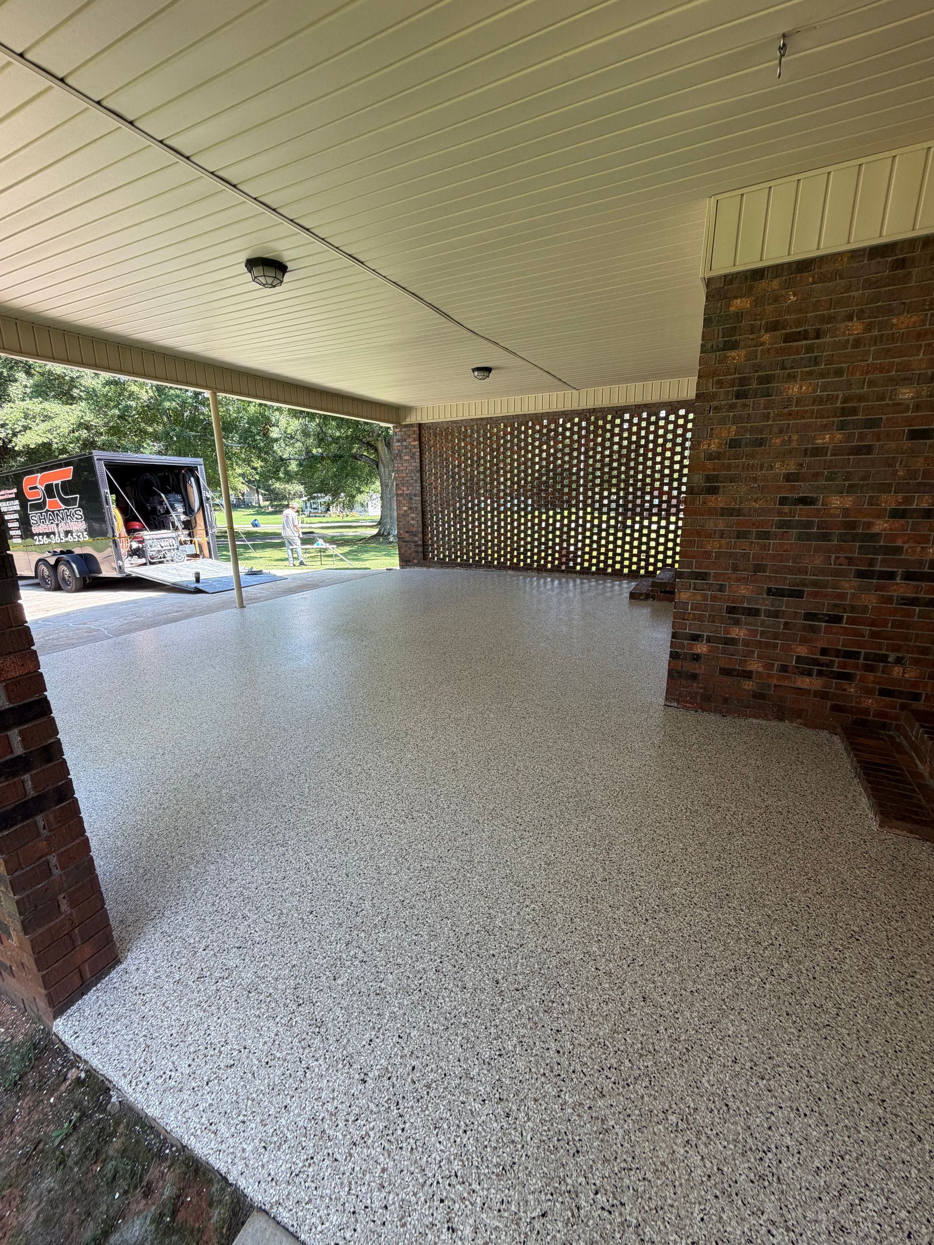 Covered brick porch with speckled floor and ceiling, opening to a leafy outdoor area.