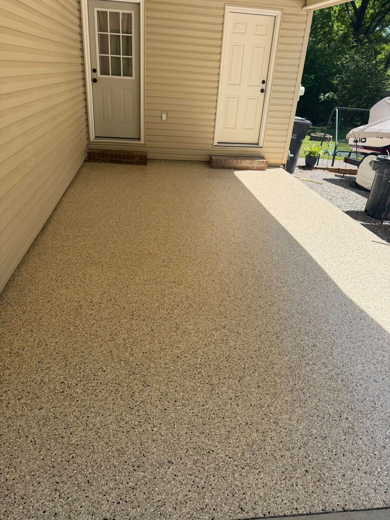 Gravel patio beside a beige house with a door, white shed, and outdoor grill in sunlight