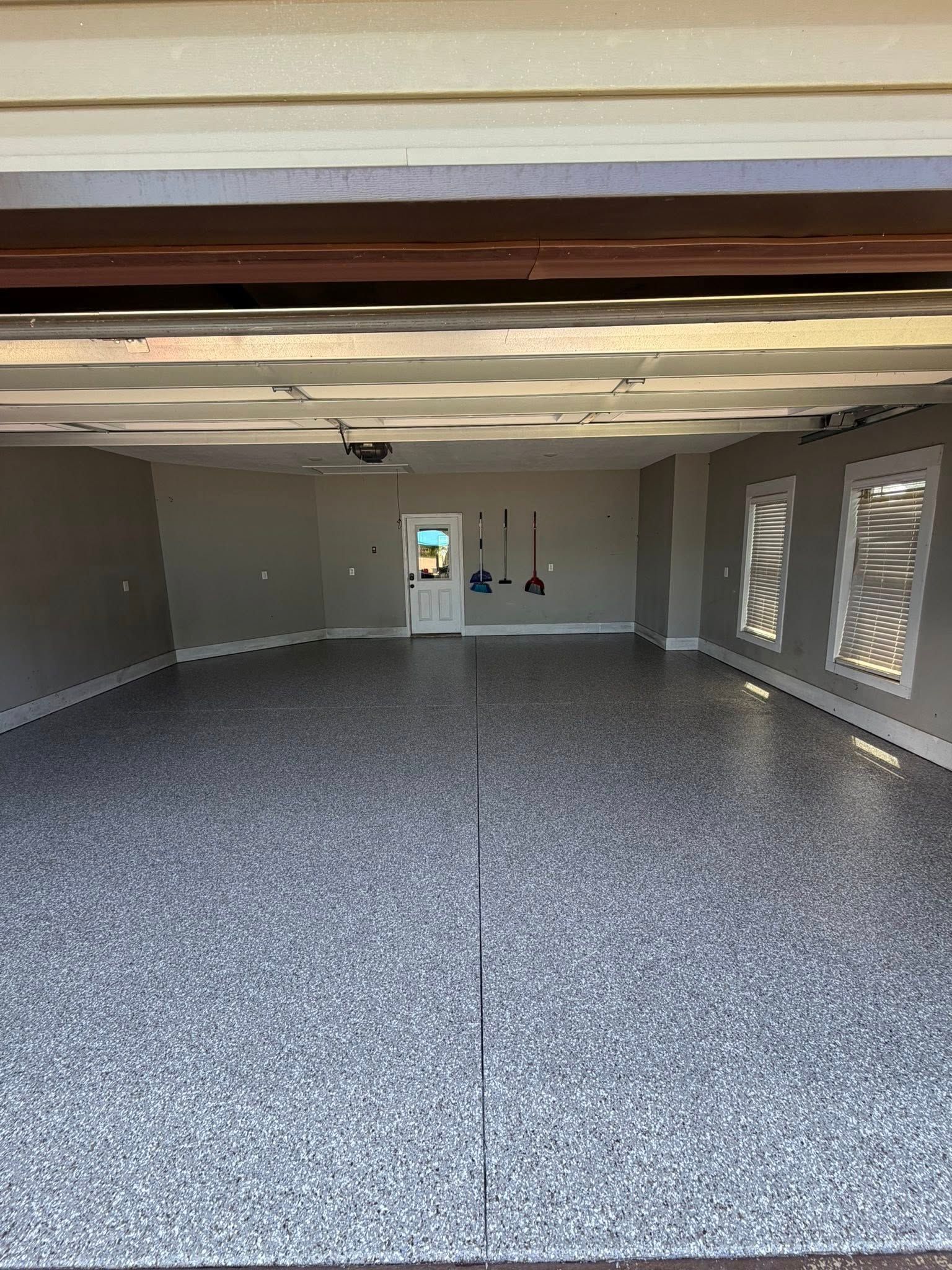 Empty gray garage with speckled floor, open ceiling beams, and a door at the far wall